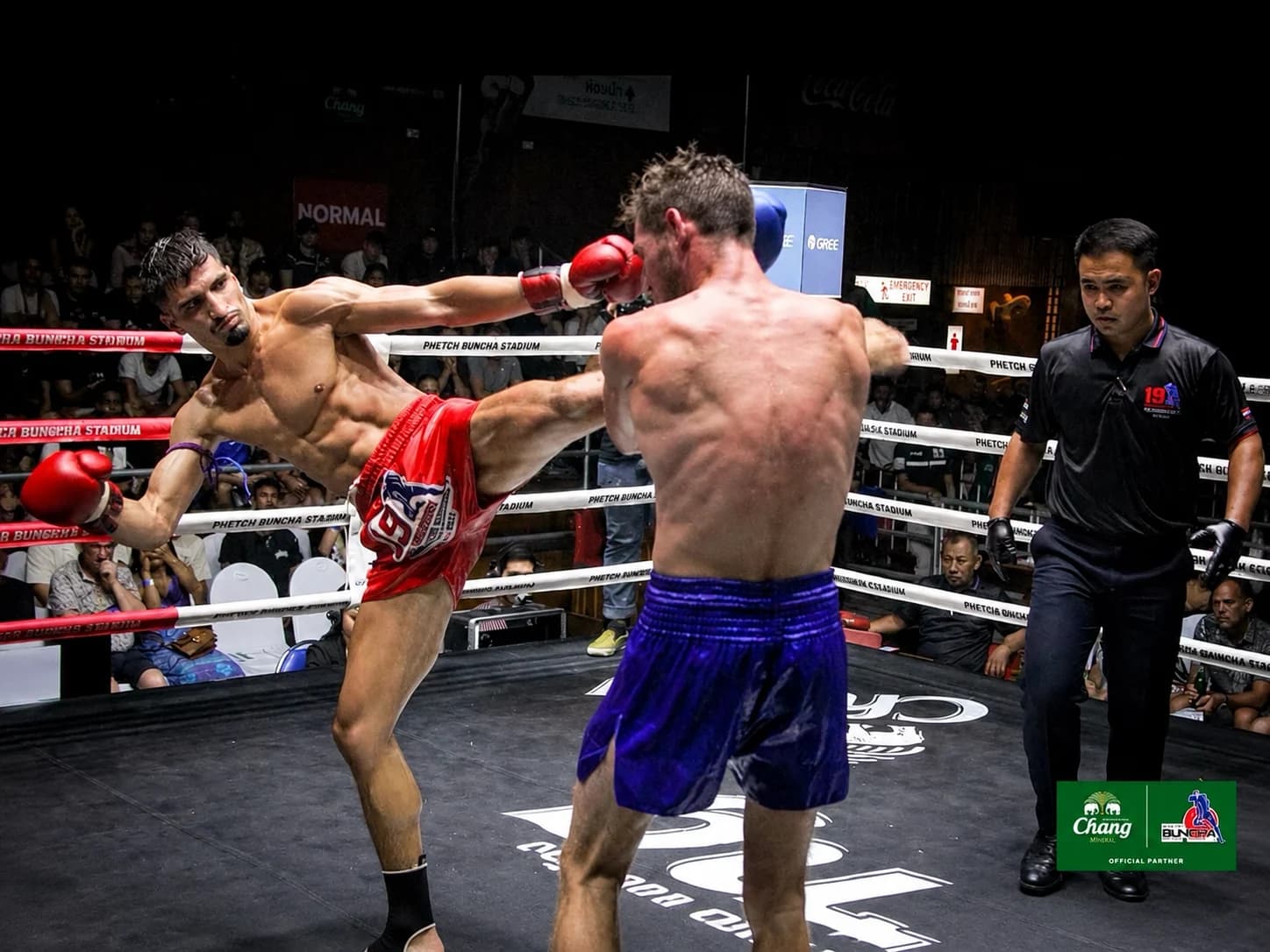 Combattant décochant un coup de pied haut lors d'un combat de Muay Thai sur le ring, public et arbitre en arrière-plan en Thaïlande