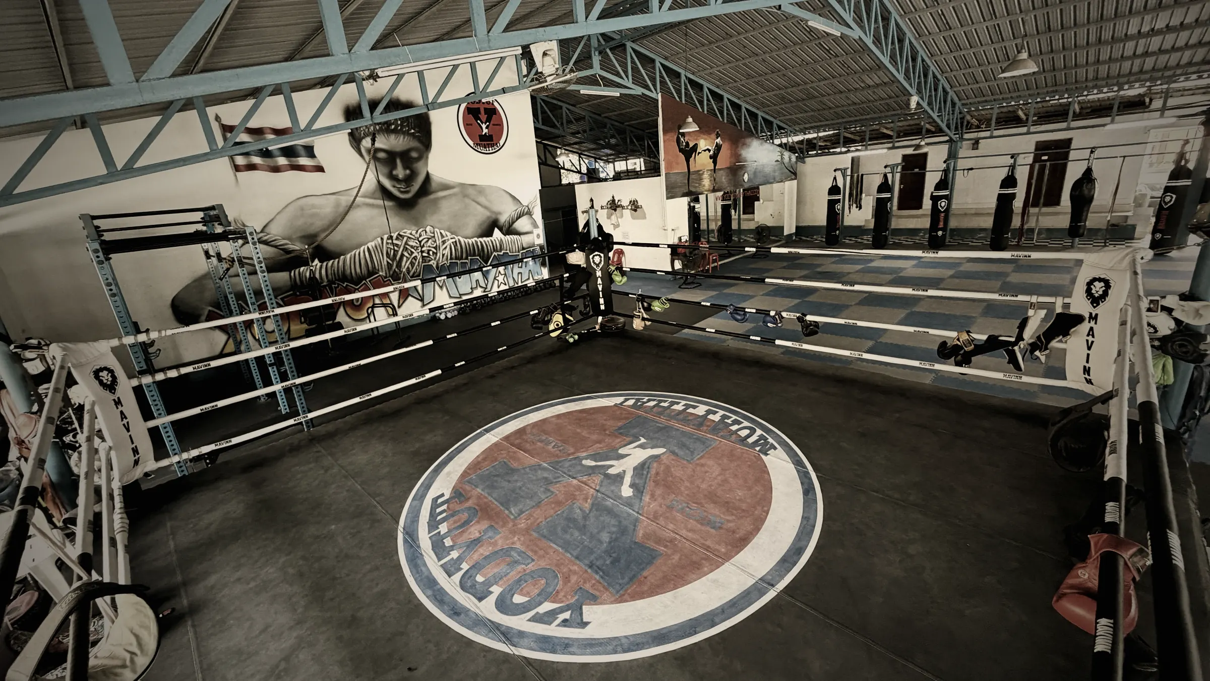 Vue d'ensemble de la salle Yodyut Muay Thai à Koh Samui : ring bleu, sacs de frappe alignés et fresque murale de combattant